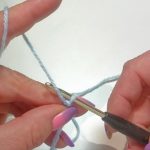 Den Arbeitsfaden über den Mittelfinger der linken Hand legen und die Startschlaufe mit Daumen und Mittelfinger festhalten. Den Faden mit der Nadel haken und...